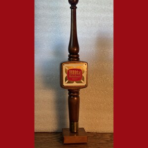 May include: A wooden beer tap handle with a gold-colored metal plate featuring the text "Blitz Weinhart Beer".