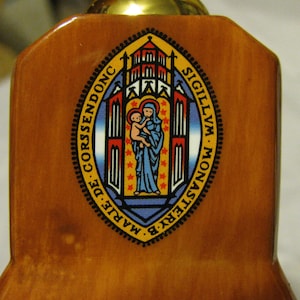 May include: A circular seal with a black border and gold lettering. The seal features a red, white, and blue design with a depiction of a building and a woman holding a child. The text on the seal reads "SIGILLVM MONASTERY DE MARIE CORSENDONC".