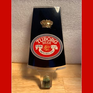 May include: A black and red plastic beer tap handle with a gold crown on top. The handle features the Tuborg Beer logo with the text "Tuborg Beer Brewed Light U.S.A. Breweries Ltd."