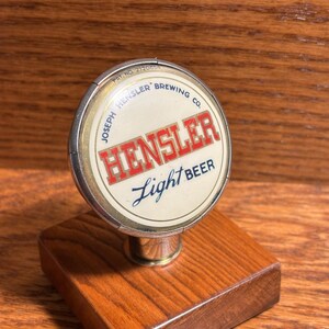 May include: A vintage Hensler Light Beer tap handle. The circular tap head features the Hensler Brewing Co. logo in red and white, with "Light Beer" in script. It is mounted on a rectangular wooden base with a polished finish.