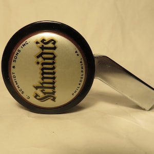 May include: A black and silver bottle opener with a round, black handle. The handle has a white and gold label with the text "C. Schmidt & Sons Inc.  Schmidts Philadelphia, Pa."
