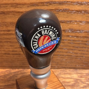 May include: A dark brown beer tap handle with the text "Galena Brewing" and "Hefe Weizen" in a circular design. The handle is mounted on a small wooden base, showcasing a classic brewery aesthetic.