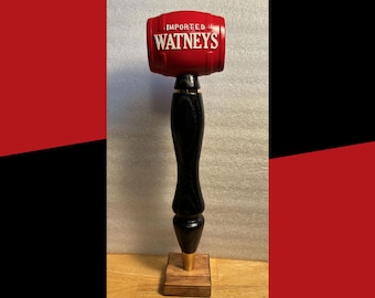 Könnte beinhalten: Ein roter, hölzerner Bierzapfhahn mit einem schwarzen Holzgriff und einer goldenen Basis. Der Zapfhahn ist fassförmig und trägt die Aufschrift "IMPORTED WATNEYS".