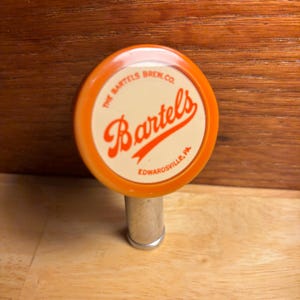 May include: A vintage beer tap handle with a circular orange and cream-colored design. The handle features the text "The Bartels Brew Co." and "Bartels" in a stylized script, with "Edwardsville, PA" below. The tap has a silver metal stem.