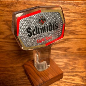 May include: A clear glass beer tap handle with a silver and red label that reads "Schmidts" and "Light Beer". The handle is mounted on a wooden base.