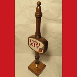 May include: A vintage wooden beer tap handle with the words "STROH LIGHT" on a rectangular label. The handle is dark brown with a square base and a rounded top. The label features red and gold accents.