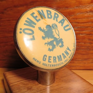 May include: Vintage Löwenbräu beer tap handle with a yellow background, a blue lion logo, and the text "Löwenbräu Germany Hans Holterbosch, Inc. NYC Bl. 51".