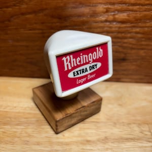 May include: A vintage beer tap handle with a white top and a red and white label that reads "Rheingold EXTRA DRY Lager Beer." The handle is mounted on a small, square wooden base.