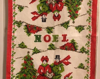 May include: A white linen tea towel with a red border and a Christmas scene. The scene features a couple singing carols, a Christmas tree, and a small child. The text "NOEL" is printed in green letters.