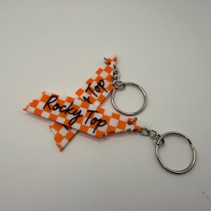 May include: Two keychains with a white and orange checkered design in the shape of a state. The words "Rocky Top" are written in black script. Each keychain has a silver-colored ring.