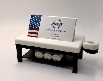 Puede incluir: Un tarjetero en forma de mesa de masaje en miniatura con superficie blanca y base negra. Sobre la mesa descansa una tarjeta de visita de U.S. Printing Press 3D Printing And Design con un gráfico de la bandera estadounidense.