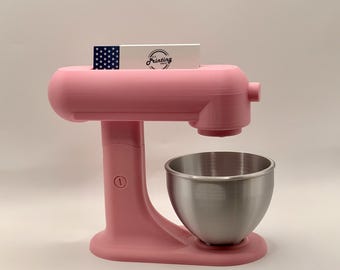 May include: A light pink stand mixer featuring a metallic silver bowl and a rectangular white card inserted into the top. The card displays a blue section with white stars on the left and the text Printing Press in black script on the right.