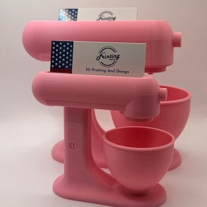 May include: Pink 3D-printed stand mixers with business cards. The mixers have a classic design, with a bowl and a head. The business cards have the text "Printing Press 3D Printing And Design" and a US flag.