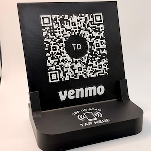 May include: Black QR code stand with the Venmo logo and the text "Tap or Scan Tap Here". The stand has a QR code with the letters "TD" in the center. The stand is designed to be used for contactless payments.