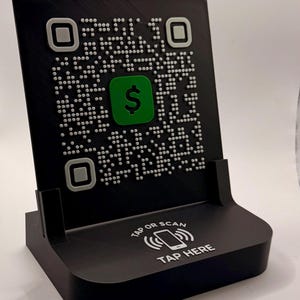 May include: Black QR code stand with a green dollar sign icon. The stand has a QR code pattern and the words "TAP OR SCAN" and "TAP HERE" are printed on the base. The stand is designed to hold a phone.