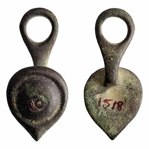 Pode incluir: Dois pingentes antigos de bronze com uma base em forma de coração e um laço circular no topo. Um pingente tem um design circular na frente, enquanto o outro tem o número "1518" inscrito na frente.