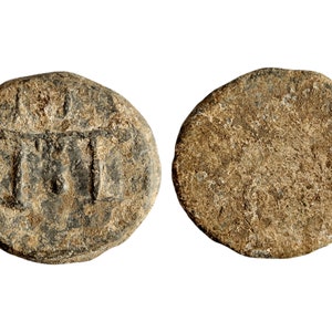 May include: A round, gray lead token with a design of three vertical lines on one side and a smooth surface on the other side.