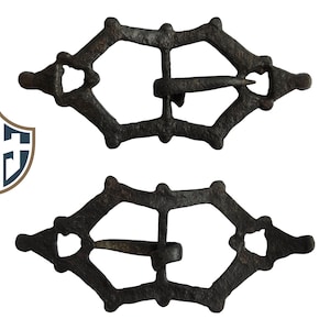 May include: Two antique, dark brown metal buckles with a diamond-shaped design. Each buckle has a central bar and a pin for fastening. The buckles are displayed against a white background.