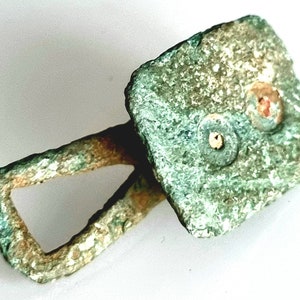 Puede incluir: Un cierre de metal cuadrado de color verde con un bucle en un lado. El cierre tiene dos agujeros circulares con un borde elevado.