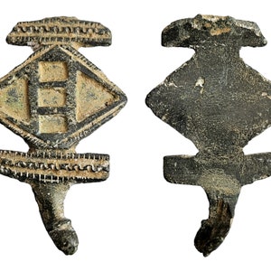 May include: A bronze pendant with a diamond-shaped design. The front of the pendant has a raised square with two horizontal lines inside. The back of the pendant is plain. The pendant has a loop at the bottom.