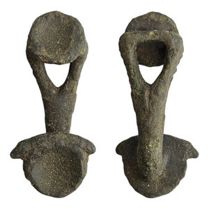 May include: A pair of antique bronze buckles with a unique design. Each buckle has a circular top with a small hole in the center and a curved, open-ended loop extending from the bottom.