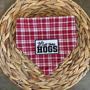 May include: A triangular pet bandana featuring a red and white plaid pattern. Centered on the fabric is a rectangular white patch with a black border, displaying the black text "Let's call those HOGS" in a mix of script and block lettering.