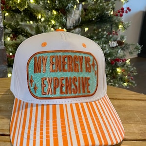 May include: White baseball cap with an orange and white striped brim. The cap features the text "MY ENERGY IS EXPENSIVE" in orange lettering on a teal background, framed by an orange border. The cap has orange button details.