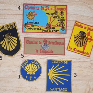 Peut inclure: Neuf patchs en tissu colorés avec des motifs de coquille Saint-Jacques et le texte "Chemins de Saint Jacques" et "Camino de Santiago".