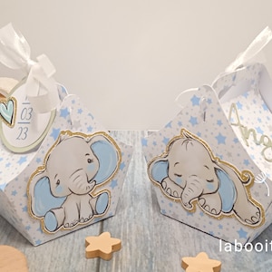 Peut inclure: Deux boîtes à dragées bleues et blanches avec un motif d'étoiles et un joli motif d'éléphant. Une boîte a une étiquette avec la date 03 23 et l'autre boîte a le nom Angelo écrit dessus.
