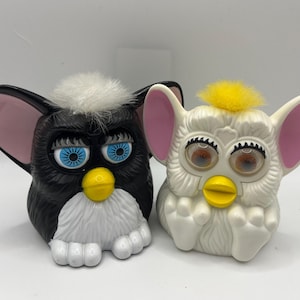 May include: Two vintage Furby toys. One is black with blue eyes, a white tuft of hair, and a yellow beak. The other is white with brown eyes, a yellow tuft of hair, and a yellow beak. Both have pink ears.