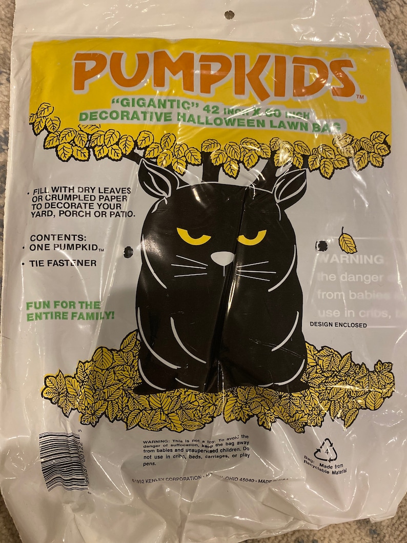 May include: A clear plastic bag containing a black cat-shaped lawn decoration for Halloween. The bag has yellow and orange text that reads "PUMPKIDS" and "GIGANTIC 42 IN x 60 INCH DECORATIVE HALLOWEEN LAWN BAG". The cat has yellow eyes.