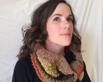 May include: A woman wearing a multi-colored crocheted cowl with brown, orange, and green tones. The cowl is made of thick yarn and has a textured, chunky look.