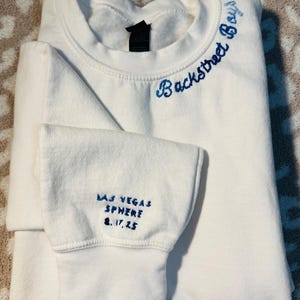 May include: A white crewneck sweatshirt with blue embroidered text. The text "Backstreet Boys" is embroidered on the upper back, and "LAS VEGAS SPHERE 8.17.25" is embroidered on the left sleeve cuff. The sweatshirt is folded.
