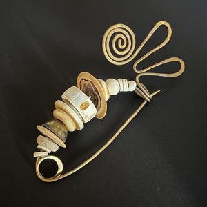 May include: A large gold-toned metal safety pin brooch featuring a decorative arrangement of various circular beads and flat discs in shades of cream, tan, and brown, with a prominent wire-formed spiral and wave design at the top of the pin.