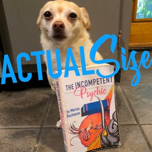 May include: A small, white dog holding a book titled "The Incompetent Psychic" by Mernie Buchanan. The book cover features a colorful illustration of a mythical creature with wings.