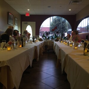 May include: A restaurant dining room with a long table set for a meal. The table is covered with a white tablecloth and set with glasses, candles, and bottles of wine. Several people are seated at the table, and the room is dimly lit.
