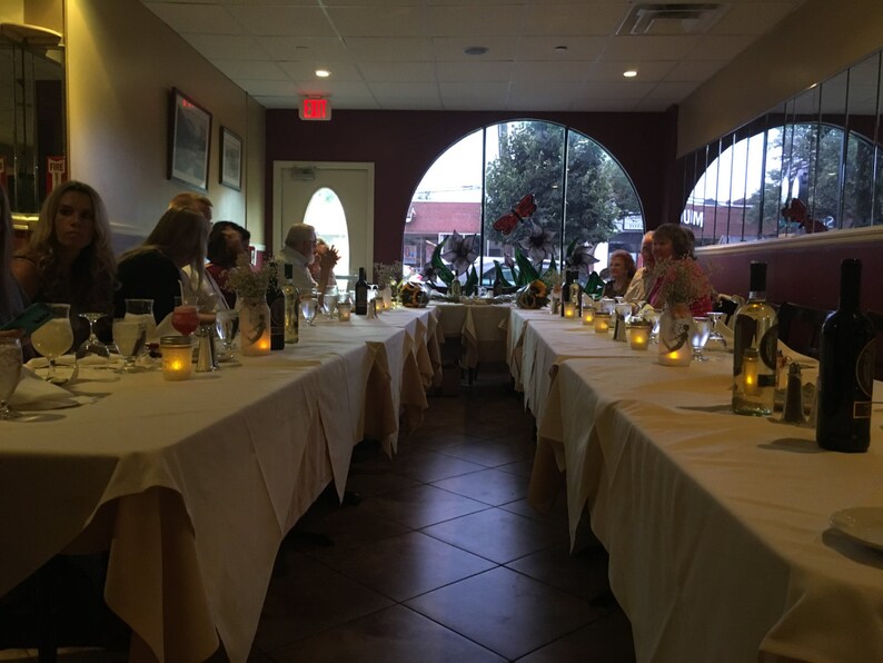 May include: A restaurant dining room with a long table set for a meal. The table is covered with a white tablecloth and set with glasses, candles, and bottles of wine. Several people are seated at the table, and the room is dimly lit.