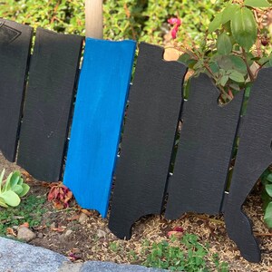 May include: A wooden United States map cutout, painted black with a blue stripe. The map is made of individual wooden slats, and is placed outdoors in a garden setting with green foliage and flowers.