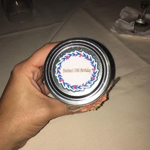 May include: A jar lid with the text "Darlene's 70th Birthday" surrounded by a blue and red floral wreath. The lid is silver and the jar is decorated with a floral design.