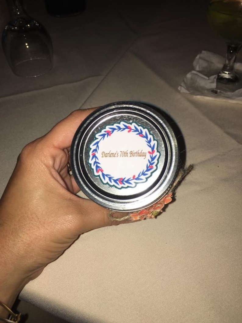May include: A jar lid with the text "Darlene's 70th Birthday" surrounded by a blue and red floral wreath. The lid is silver and the jar is decorated with a floral design.