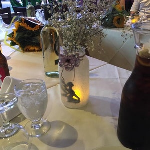 May include: A table setting with a lit mason jar candle holder featuring a silhouette of a fairy. The jar is decorated with a purple flower and filled with baby's breath. Other items include wine bottles, glasses, and a pitcher of a dark liquid.