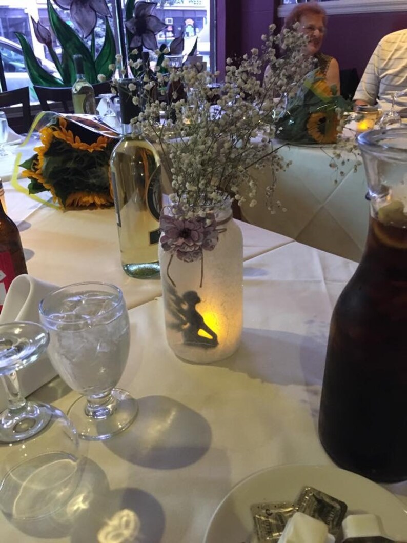 May include: A table setting with a lit mason jar candle holder featuring a silhouette of a fairy. The jar is decorated with a purple flower and filled with baby's breath. Other items include wine bottles, glasses, and a pitcher of a dark liquid.