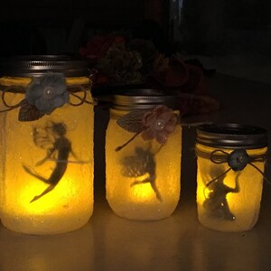 May include: Three illuminated mason jars with fairy silhouettes inside. Each jar is decorated with a flower and twine. The jars are different sizes, creating a whimsical, glowing effect.