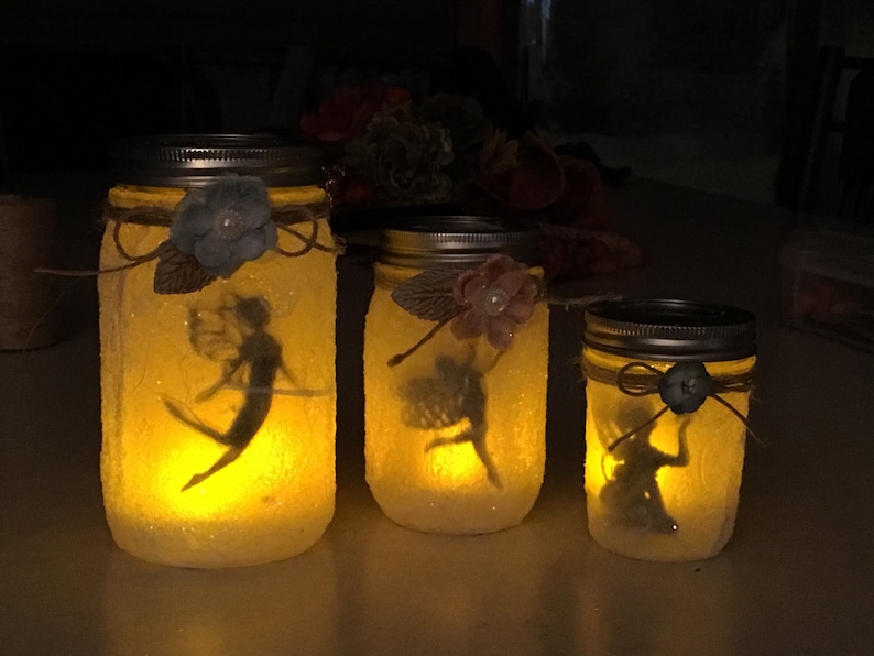 May include: Three illuminated mason jars with fairy silhouettes inside. Each jar is decorated with a flower and twine. The jars are different sizes, creating a whimsical, glowing effect.