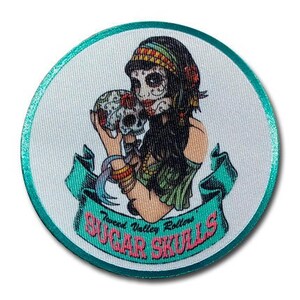 May include: A round, teal-coloured patch with a white background. The patch features a colourful illustration of a woman with a skull in her hand. The woman has long black hair, a colourful headband, and a green and white dress. The text "Tweed Valley Rollers Sugar Skulls" is printed in pink and white on the patch.