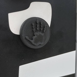 May include: A black handprint embossed on a round, black button. The button is attached to a black and white striped surface.