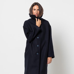 Puede incluir: Un abrigo largo azul marino con cuello alto y cierre de botones. El abrigo llega a la rodilla y parece estar hecho de un material suave y cálido. La modelo lleva vaqueros azules y zapatos oscuros.