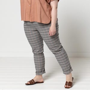 Könnte beinhalten: Eine Frau trägt ein kurzärmeliges, knöpfbares Hemd in Rostbraun und eine karierte Hose. Die Hose ist schwarz, weiß und beige. Sie trägt auch braune Sandalen. Das Outfit ist lässig und stilvoll.
