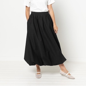 May include: A woman wearing a white t-shirt and a black midi skirt with pockets. She is wearing light-colored flats. The skirt has a full, gathered design. The background is a plain white wall and floor.
