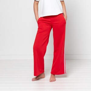 May include: A person wearing a white t-shirt with short sleeves and red sweatpants. The sweatpants have a white stripe down the side. The person is standing in front of a white background.
